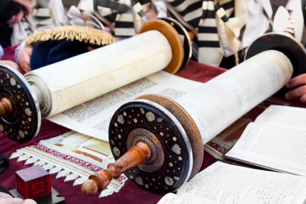 Open Torah scrolls on a table with a person reading, surrounded by traditional garments.