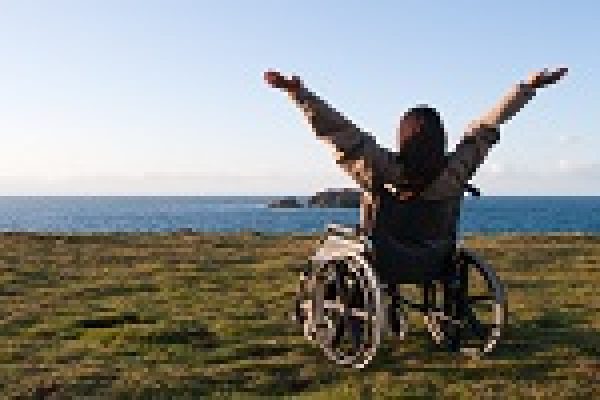 Person in wheelchair with arms raised, overlooking the sea on a grassy cliff under a clear blue sky.