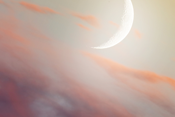 A crescent moon in a soft, pastel sky with pink and orange clouds at dusk or dawn.