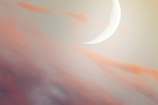 Crescent moon in a softly lit sky with clouds in pink and orange hues.