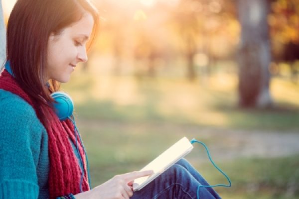 Woman sitting outdoors, reading on a tablet with headphones around her neck, in a sunlit park.