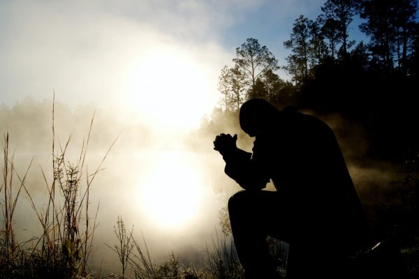 Silhouette of a person kneeling and praying by a misty lake, with trees and the sun in the background.