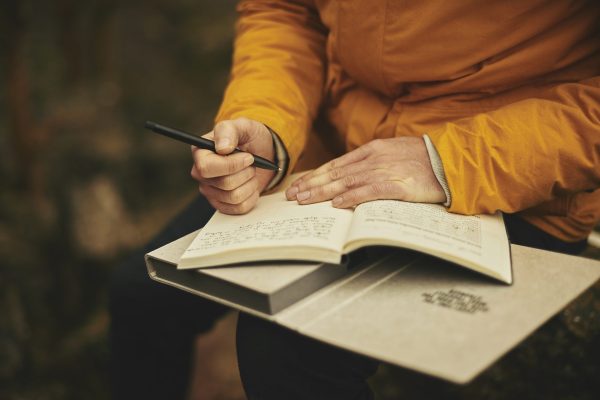 Person in orange jacket writing in a journal while sitting outdoors.