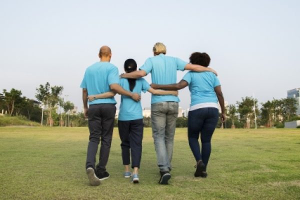 Four people in blue shirts walk arm-in-arm on a grassy field, facing away from the camera.