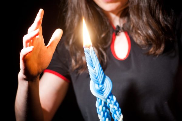A woman holds a lit blue braided candle, raising her hand toward its flame against a dark background.