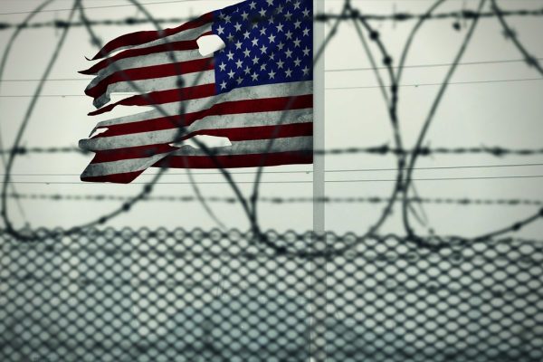 a barbed wire fence stands in front of a torn American flag waving against a gray sky