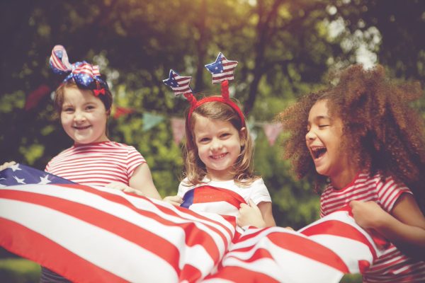 Three children smiling, wearing striped shirts and holding an American flag, with trees in the background.