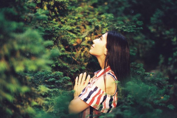 brown skinned woman with long dark hair in forest, hands pressed together, looking up, with a look of wonder or gratitude
