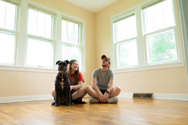 Couple sits on a wooden floor in a bright room with a black dog, smiling near large windows.