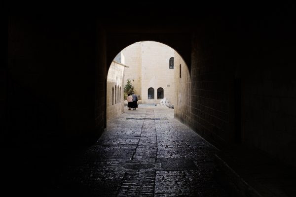 A person walks through a dimly lit stone tunnel towards a sunlit courtyard.