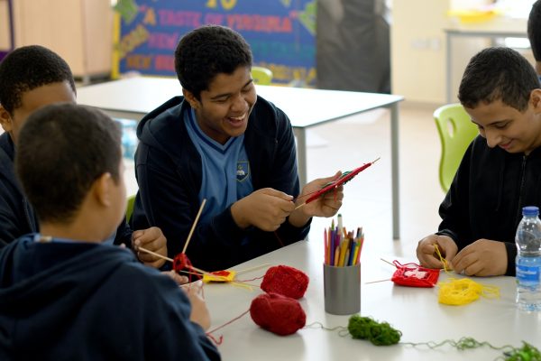 Group of boys smiling and knitting with colorful yarn at a classroom table.