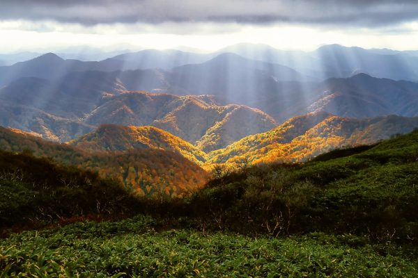 Sunbeams shine through clouds onto green and autumn-colored hills and mountains in a scenic landscape.