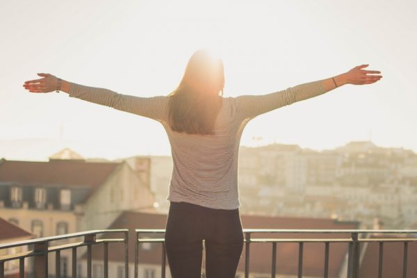 Person with arms outstretched stands on a balcony overlooking a sunny cityscape.