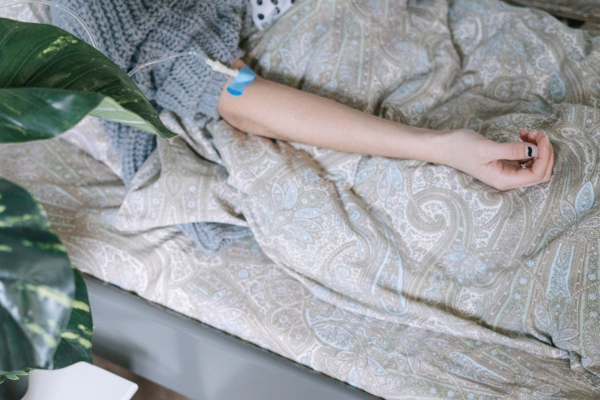 A person lying in bed with an IV in their arm, covered by a patterned blanket, next to a leafy green plant.