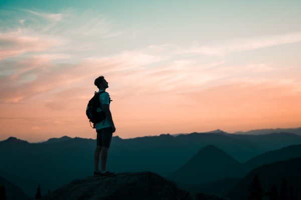 A hiker with a backpack stands on a rock, gazing at a sunset over mountains.