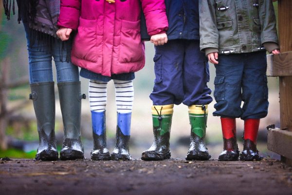 Four children in colorful rain boots stand on a muddy path, wearing cozy winter jackets.