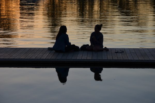 Silhouettes of two people sitting on a dock by a calm lake during sunset, reflecting on the water.