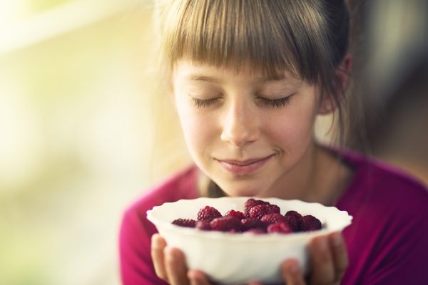 Girl with closed eyes holds and smells a bowl of raspberries, wearing a pink shirt and smiling softly.