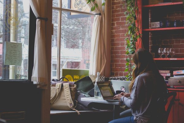 Person typing on a laptop in a cozy cafe with large windows and plants.