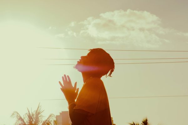 A person stands silhouetted against a bright sunset sky with palms, gazing upwards with hands in a prayer position.