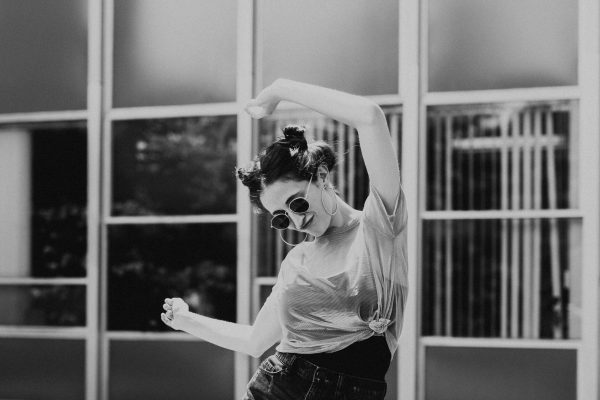 black and white photo of woman dancing, arms in the air, head down. She is wearing sunglasses and has her hair up in buns.