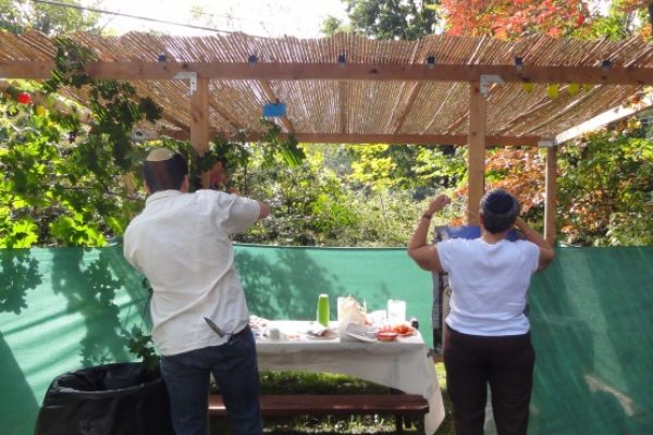 Two people decorating an outdoor sukkah with greenery, under a wooden roof, with a table of supplies nearby.