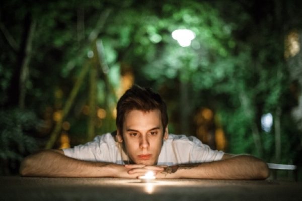 A person in a white shirt leans on a surface, gazing at a glowing light, with a blurred green background.