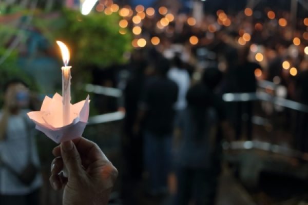Hand holding a lit candle with a paper holder in a crowd of people, with blurred lights in the background.