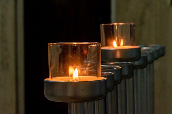 Two lit votive candles in glass holders, placed in a row on metallic stands in a dimly lit setting.