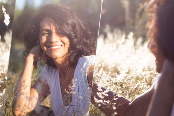 Woman smiling at her reflection in a mirror placed on grass with white flowers.