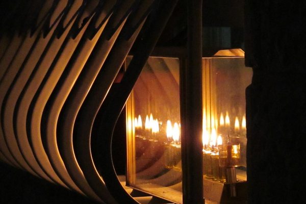 A close-up of candles lit in a reflective menorah, casting warm light and shadows through a geometric pattern.