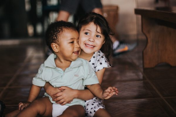 Two young children sitting on the floor, with one hugging the other and smiling.