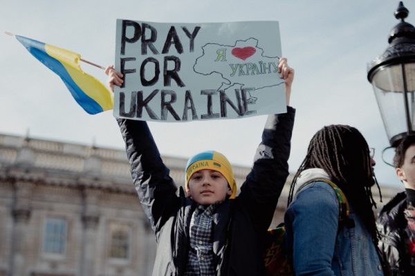 Person holding Pray for Ukraine sign at a rally, with a Ukrainian flag nearby and people in the background.