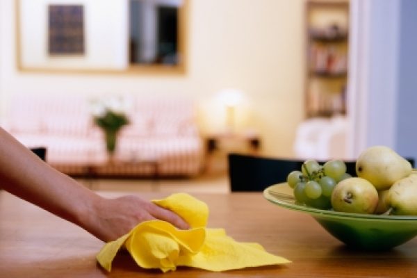 Hand wiping wooden table with yellow cloth, fruit bowl in foreground, cozy living room in background.