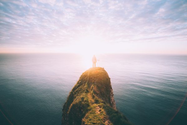 large rock jutting into the water. A person stands on the rock's edge looking out into a bright, cloudy horizon