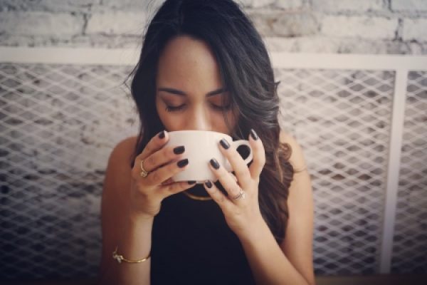 A woman with dark hair enjoys a hot drink from a white cup, sitting in a cozy setting.