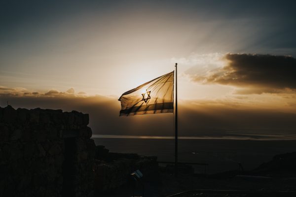 Flag waving in front of a dramatic sunset, casting a silhouette over a rocky landscape and cloudy sky.