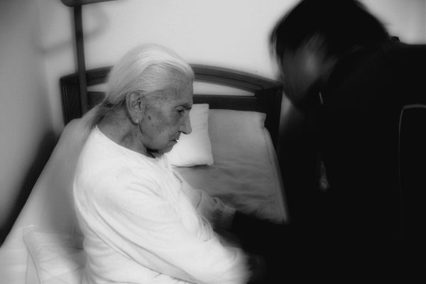 Elderly person sitting on a bed, with another person beside them in a dimly lit room.