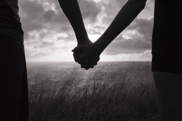Two people holding hands, silhouetted against a cloudy sky and grassy landscape near the water.