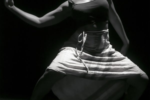 a black and white photo of a dancer with short hair wearing a long skirt with bent knees, face angled upward, and arm extending to the side