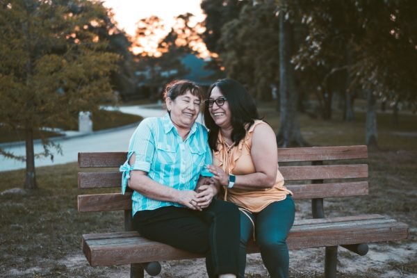 Two women sitting on a park bench, smiling and laughing, with trees in the background at sunset.