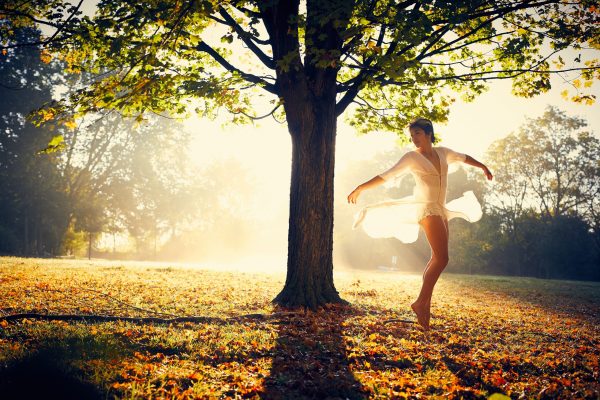 tree in the center of image with light shining through, woman dancing to the right, wearing a gauzy white dress that's blowing as she dances with her hands outstretched. she has brown hair and light or olive skin.