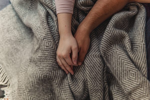 Two hands gently holding each other resting on a patterned gray blanket.