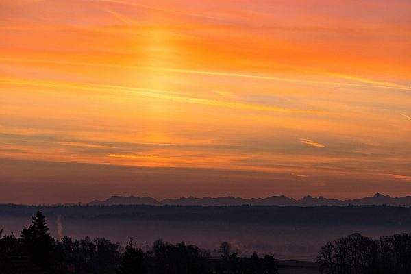 Orange and pink sunset over distant mountain silhouette with a misty foreground of trees.