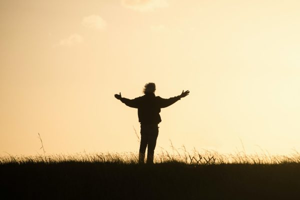 Silhouette of a person with outstretched arms standing in a field during sunset, with grass and sky in the background.