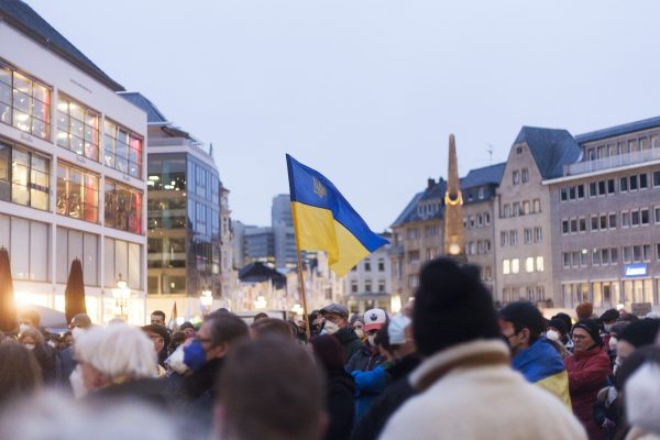 Crowd gathered in a city square with a Ukrainian flag prominently raised in the center.