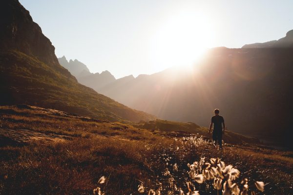 Person walking in a sunlit mountainous landscape with a bright, setting sun and clear sky.