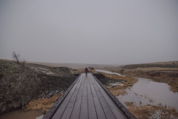 A person walks on a wooden bridge over a misty river in a foggy landscape.