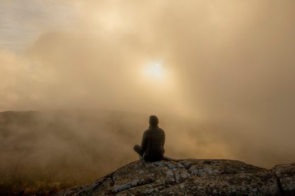 Person sitting on a rock, gazing at a misty sunrise over a foggy landscape.