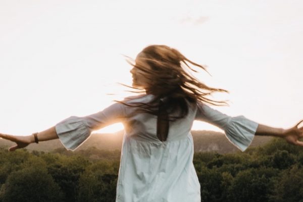 A woman with long hair spins joyfully outdoors, arms outstretched, with a serene landscape and sunset in the background.